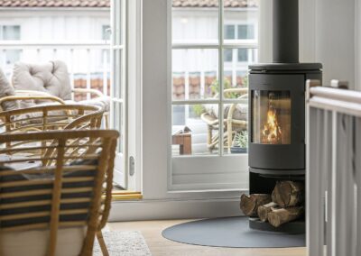 Brændeovn i sommerhus med ild i kaminen, stablet brænde og udgang til terrasse med loungemøbler, fotograferet i Danmark
