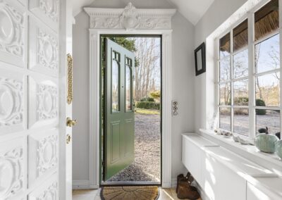 Entré i sommerhus med udsmykket hvid dør, åben grøn hoveddør og udsigt til naturgrund, fotograferet til sommerhusmarkedsføring i Vestsjælland