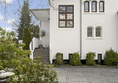 Facade set fra indkørsel med hvid mur, trappe og beplantning, fotograferet til professionel ejendomspræsentation i Danmark